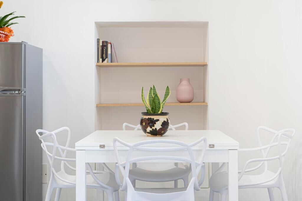 Une table à manger blanche, accompagnée de quatre chaises blanches, d’une plante en pot et d’un vase rose posé sur une étagère.