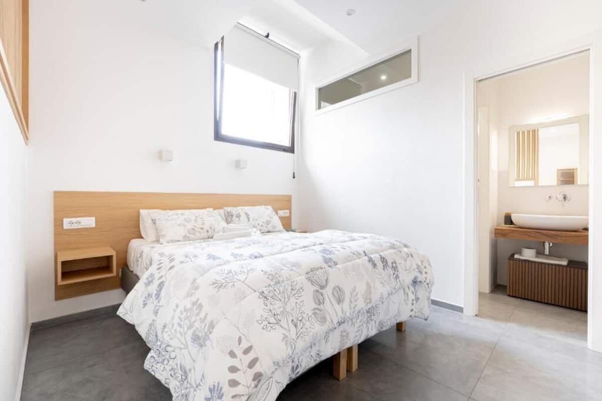 Een moderne slaapkamer met een bed, nachtkastjes, een inbouwkast, een raam en een badkamer met een wastafel en spiegel.