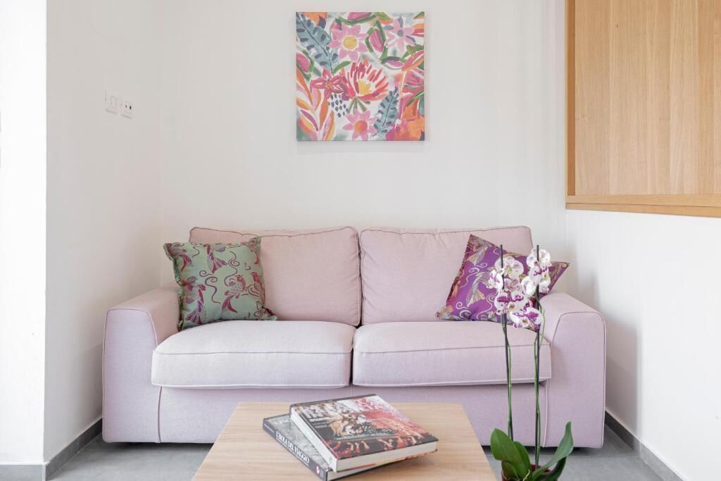 A light pink sofa with floral cushions, a small wooden table with books, and a colorful abstract painting on the wall.