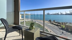 A balcony with a view of a port, a chair, a small table with a plant, and a clear blue sky.