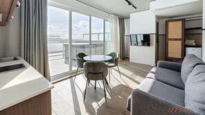 A modern living room with a sofa, a dining table with chairs, and large windows offering a view of the sea.