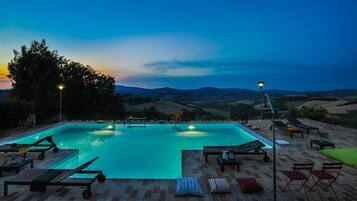 A rectangular swimming pool with lounge chairs and tables, surrounded by a brick patio, set against a backdrop of hills and a twilight sky.