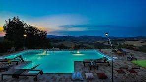Piscina rectangular con sillas y mesas de descanso, rodeada por un patio de ladrillo, con colinas y un cielo crepuscular al fondo.