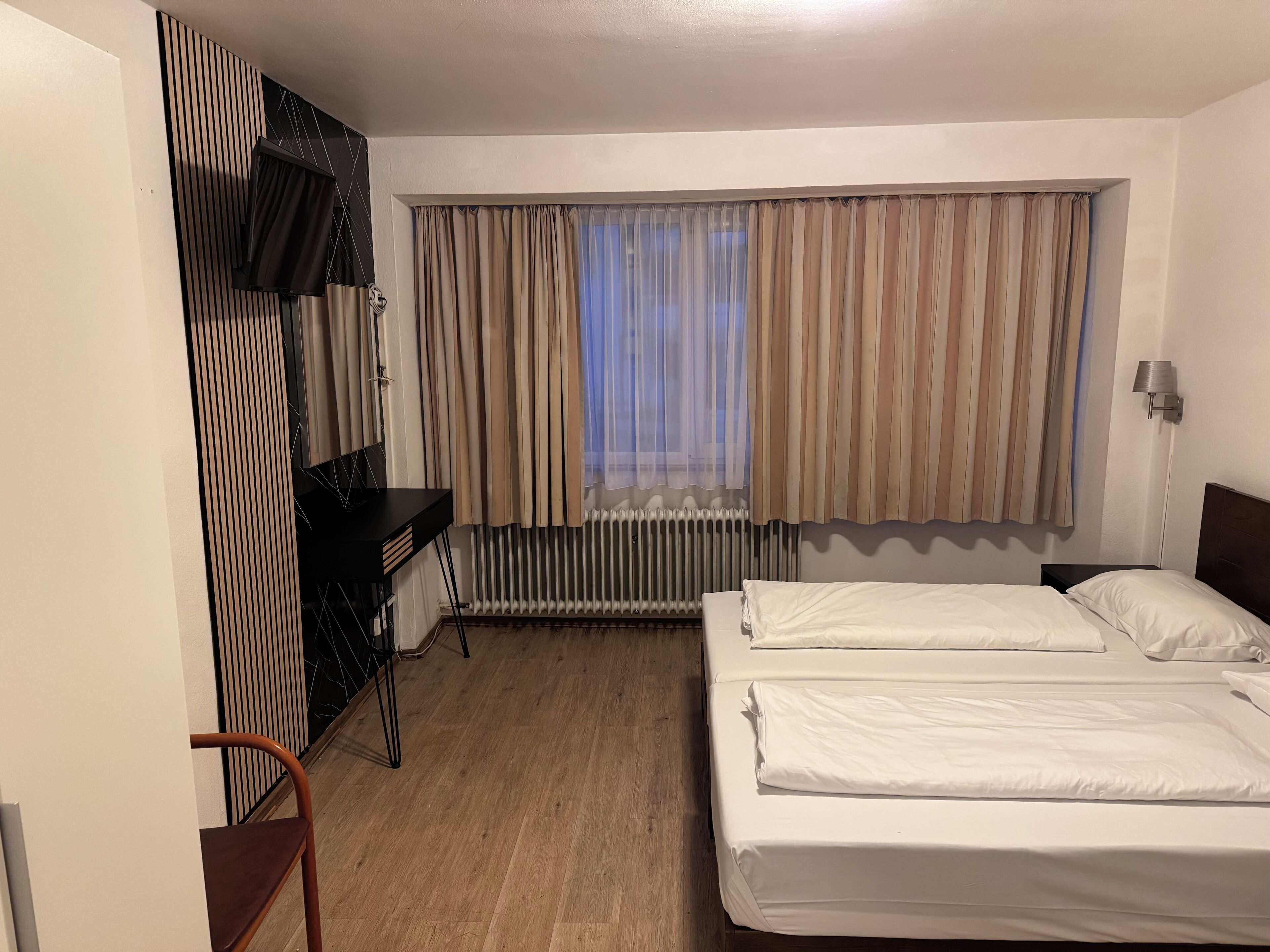 Habitación de hotel con dos camas, un escritorio, una silla, un radiador y una ventana con cortinas.