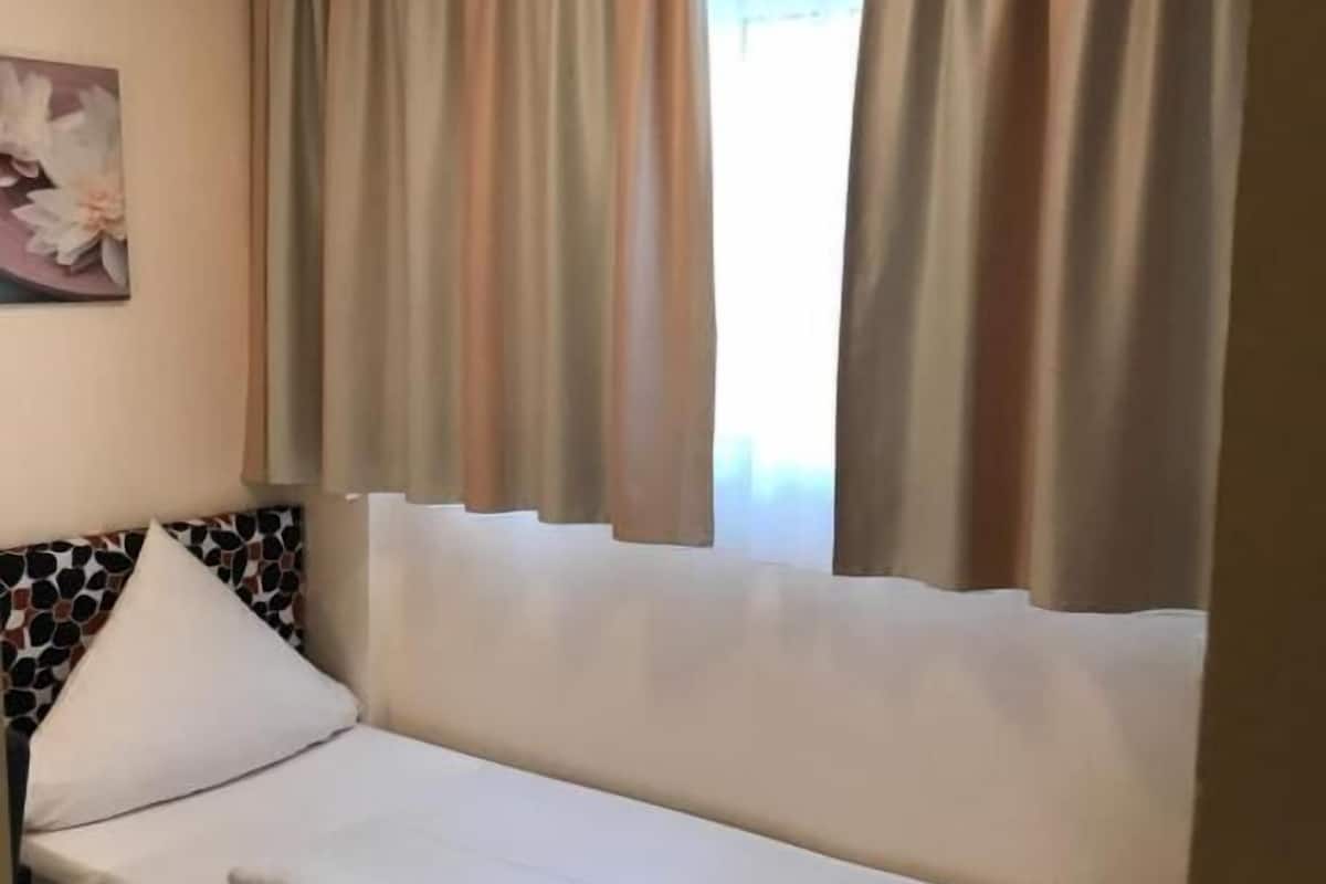 A single bed with a headboard featuring a mosaic pattern, beige curtains, and a painting of flowers on the wall.