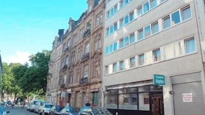 Egy utcai panoráma, amelyben egy sorban épületek láthatók, köztük egy kétszintes épület, amelyen a „Pension Alpha” feliratú tábla található, valamint egy háromszintes épület, melynek számos ablaka van.