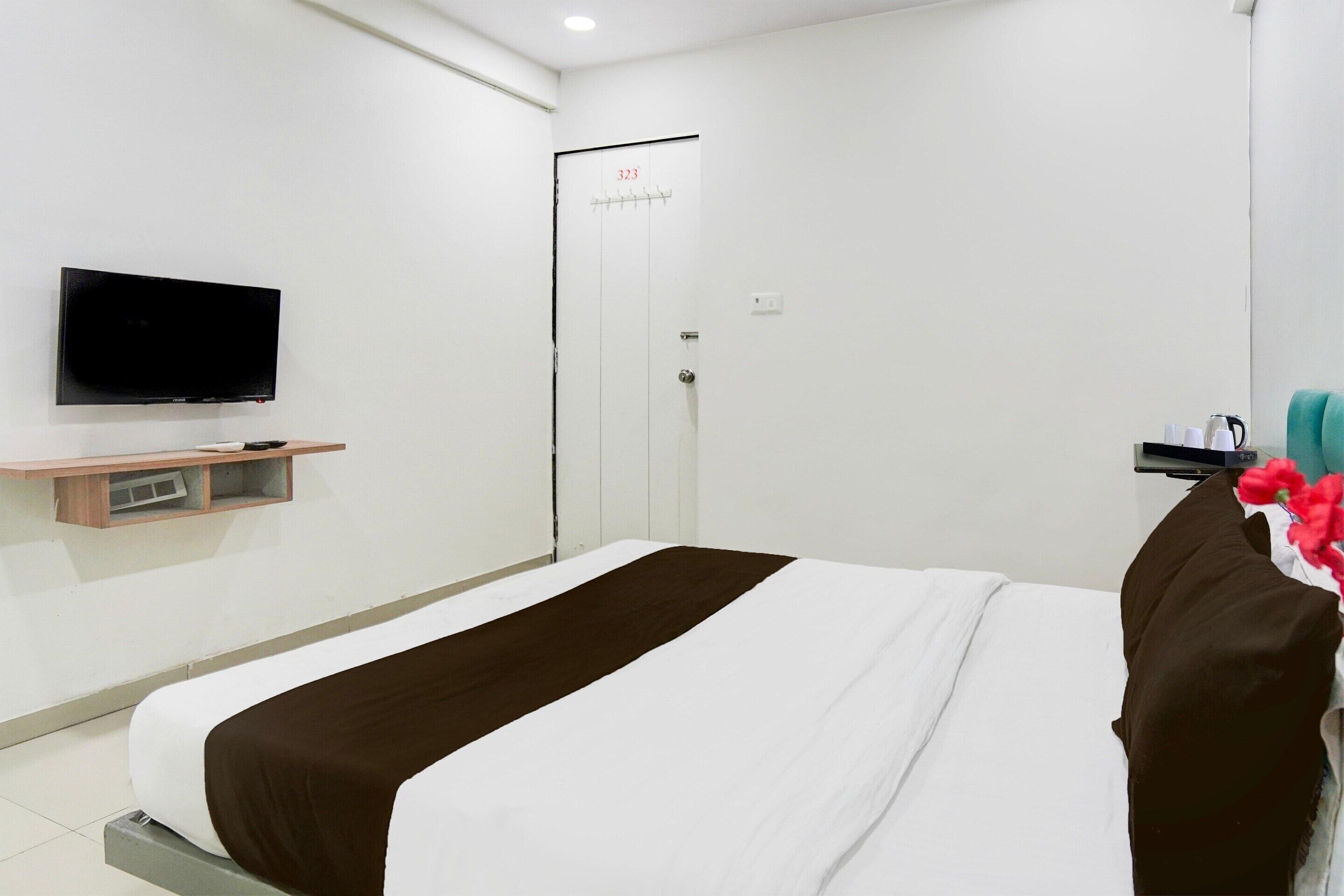A hotel room with a bed, a television mounted on the wall, a door with a number, and a small shelf with items.