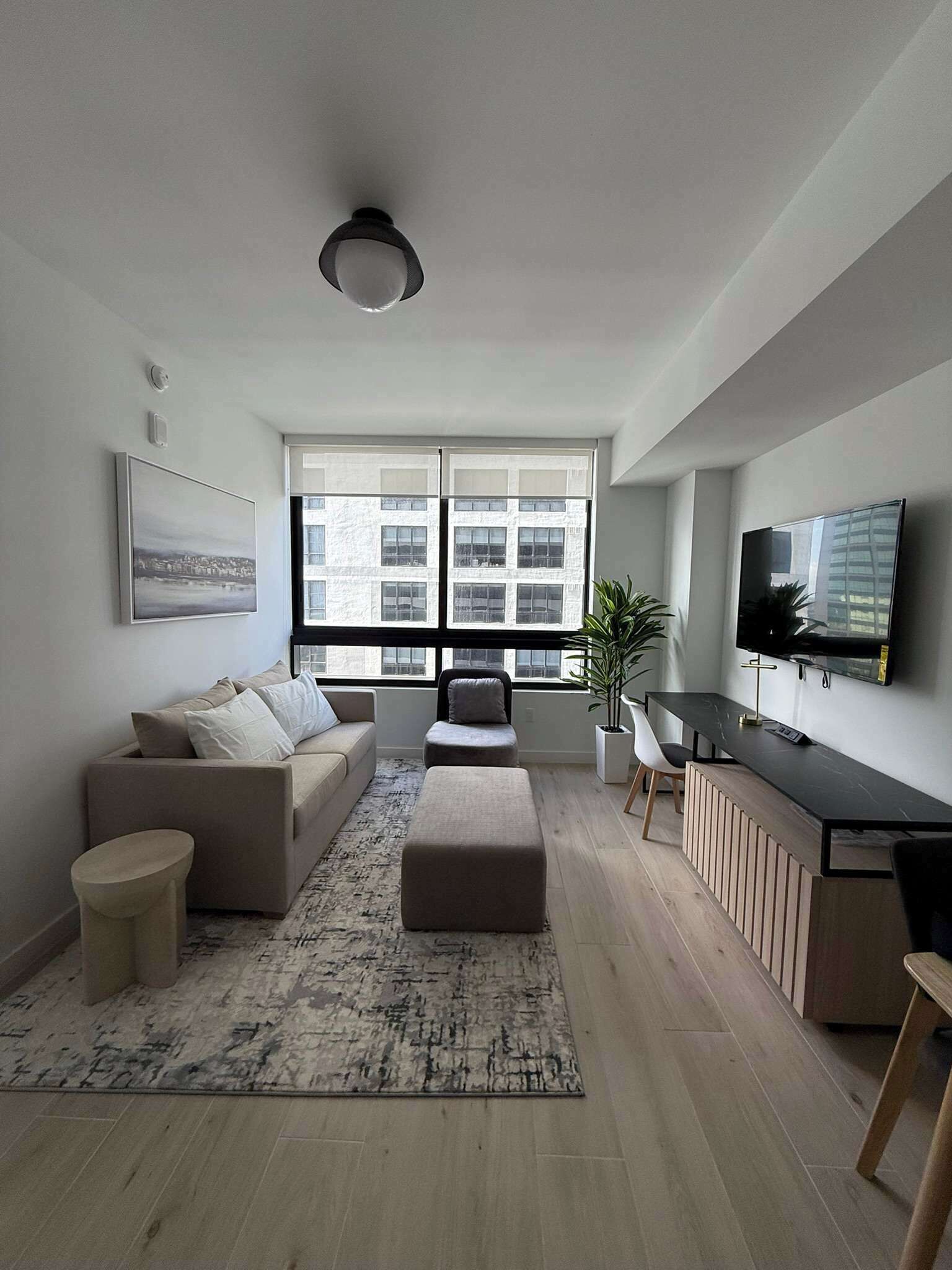A modern living room with a sofa, ottoman, and a small table. There is a large window with a view of buildings, a flat-screen TV mounted on the wall, and a potted plant.