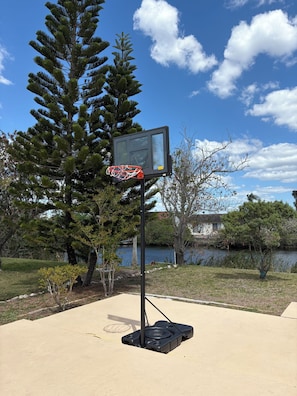 Ein Basketballplatz im Freien mit Korb, umgeben von Bäumen und einem Gewässer.
