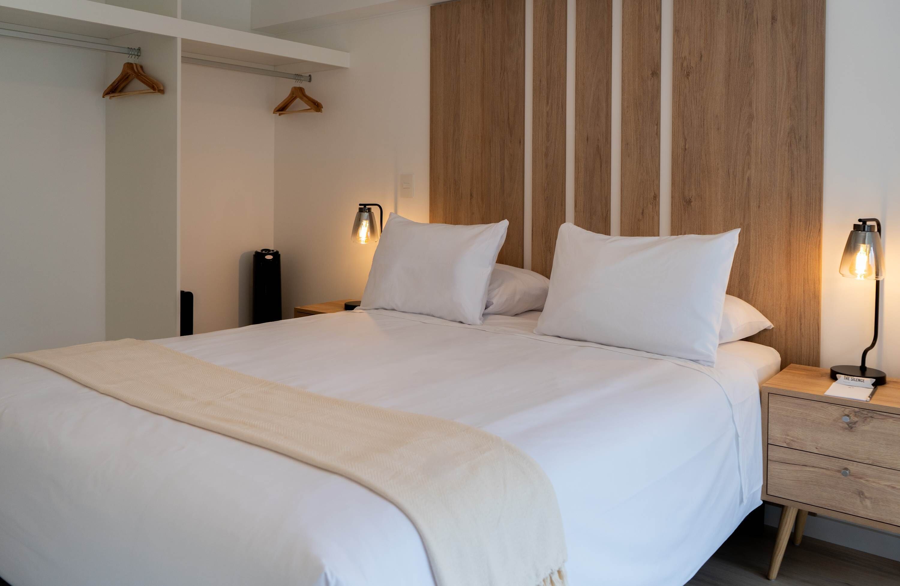A neatly made bed with white linens and a beige throw blanket. There are two bedside tables with lamps and a wooden headboard.