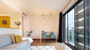A modern living room with a sofa, a dining table with chairs, a large window with a view, and a hanging light fixture. - Enjoy Apartments Maria (Barcelona)