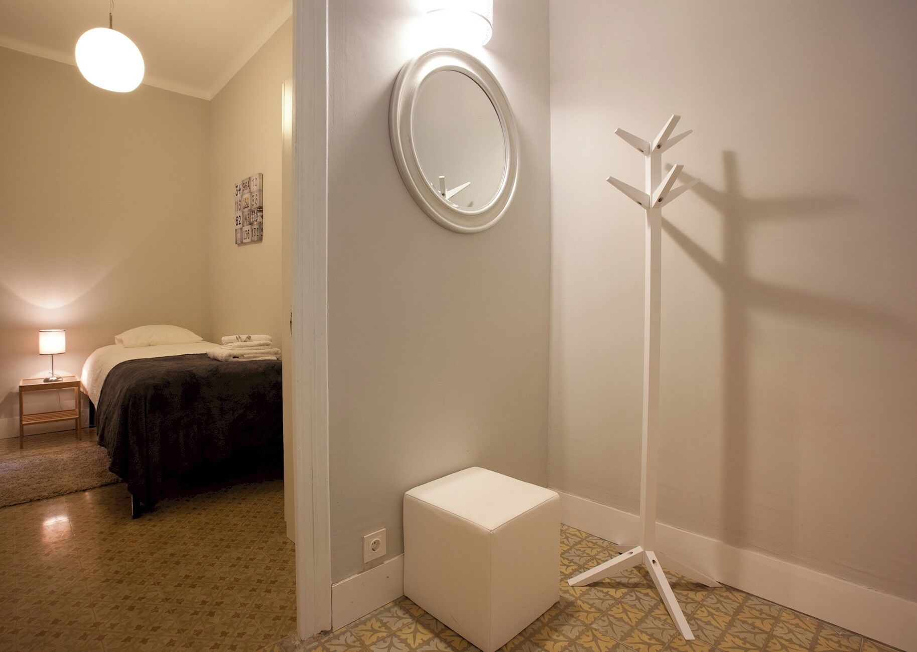 A hotel room with a bed, a bedside table with a lamp, a small white stool, a coat rack, and a mirror on the wall.