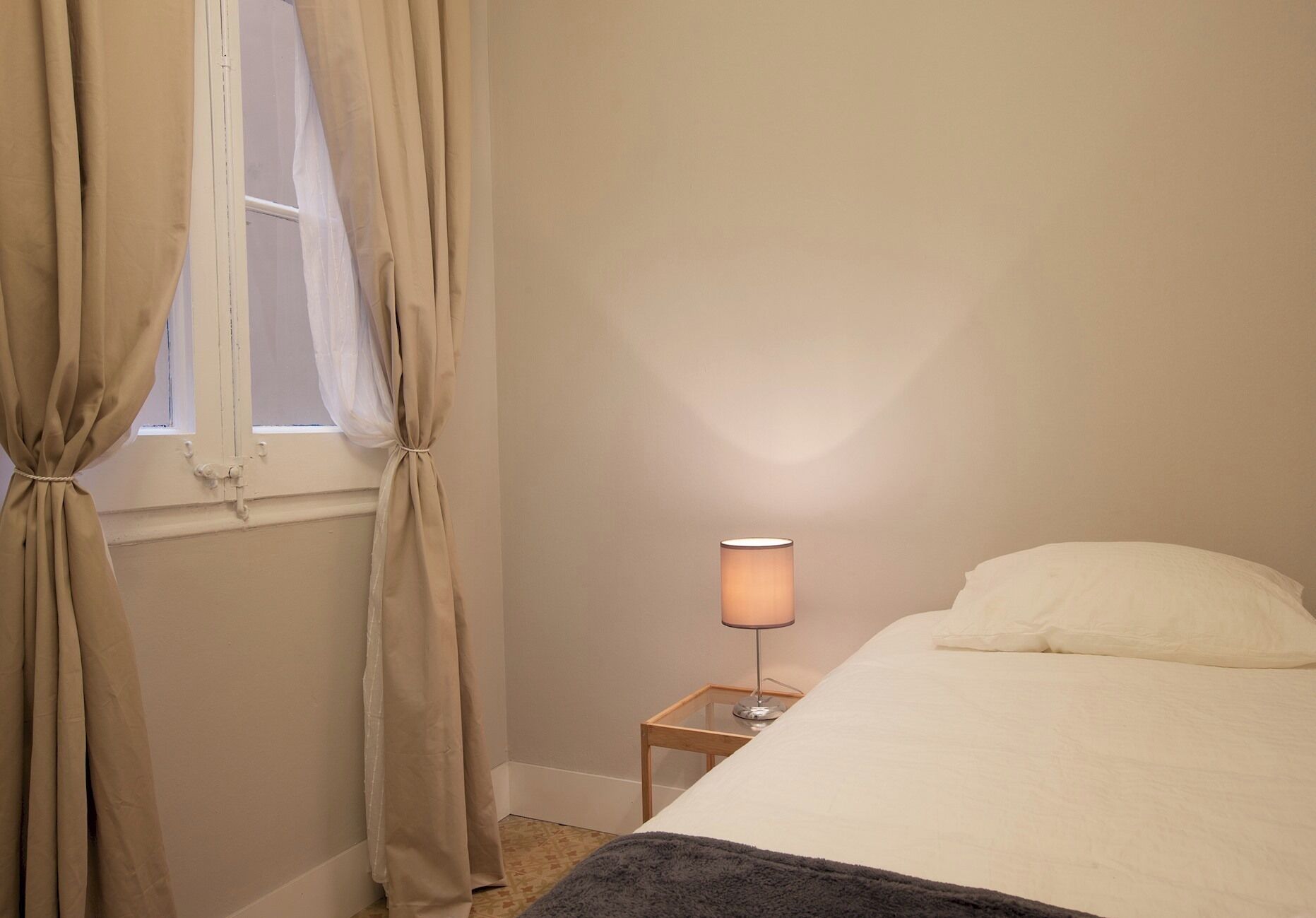 Un dormitorio con una cama, una lámpara de noche y cortinas color beige.