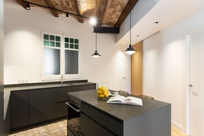 A modern kitchen with a central island, a window with frosted glass, pendant lighting, and a door to the right. - Enjoy Apartments La Riera One (Barcelona)
