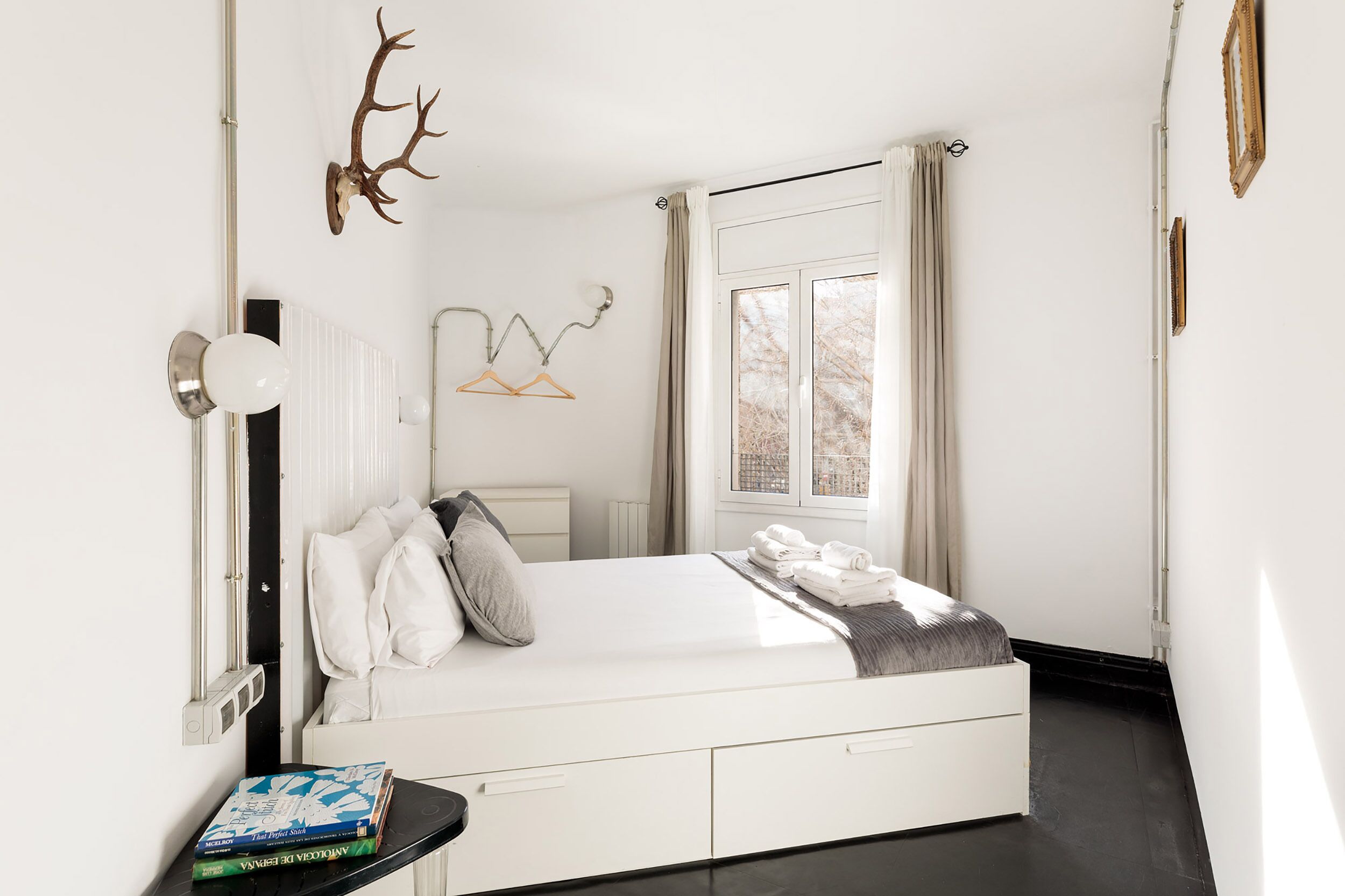 Une chambre moderne avec un lit blanc, une table de chevet avec des livres, une tête de cerf accrochée au mur et une fenêtre avec des rideaux.