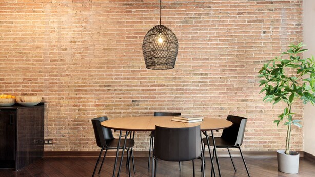 A dining area with a brick wall, a round wooden table, four black chairs, a hanging light fixture, and a potted plant. - Enjoy Apartments Urban Retreat Valencia (Barcelona)