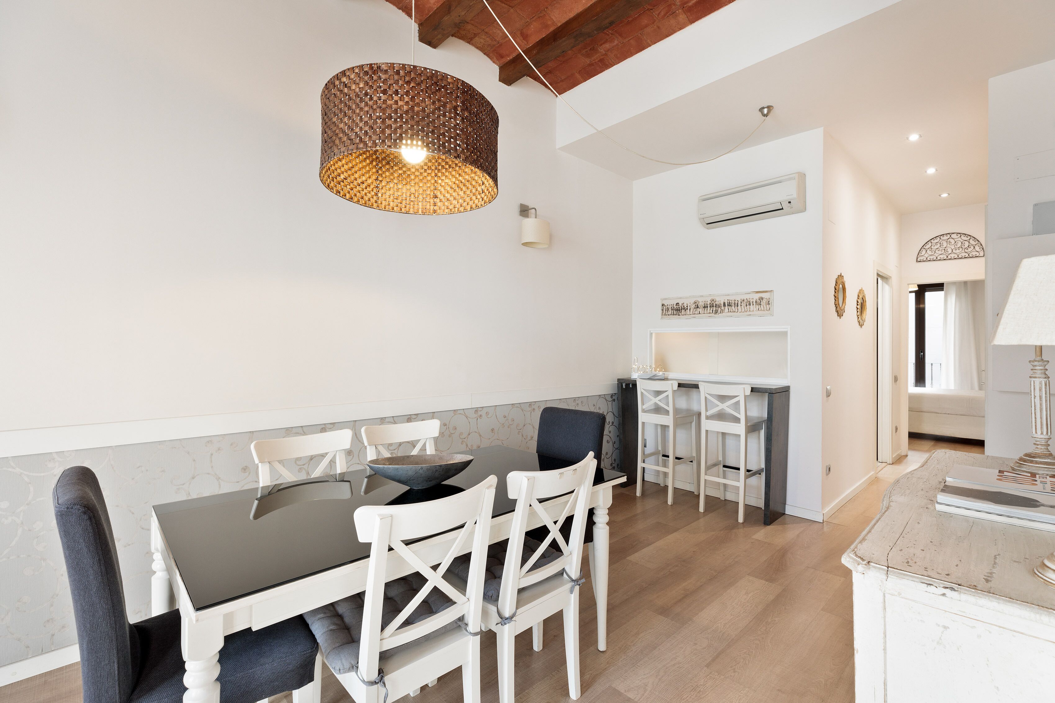 A dining area with a black table, white chairs, a woven pendant light, and a white wall with a decorative shelf.