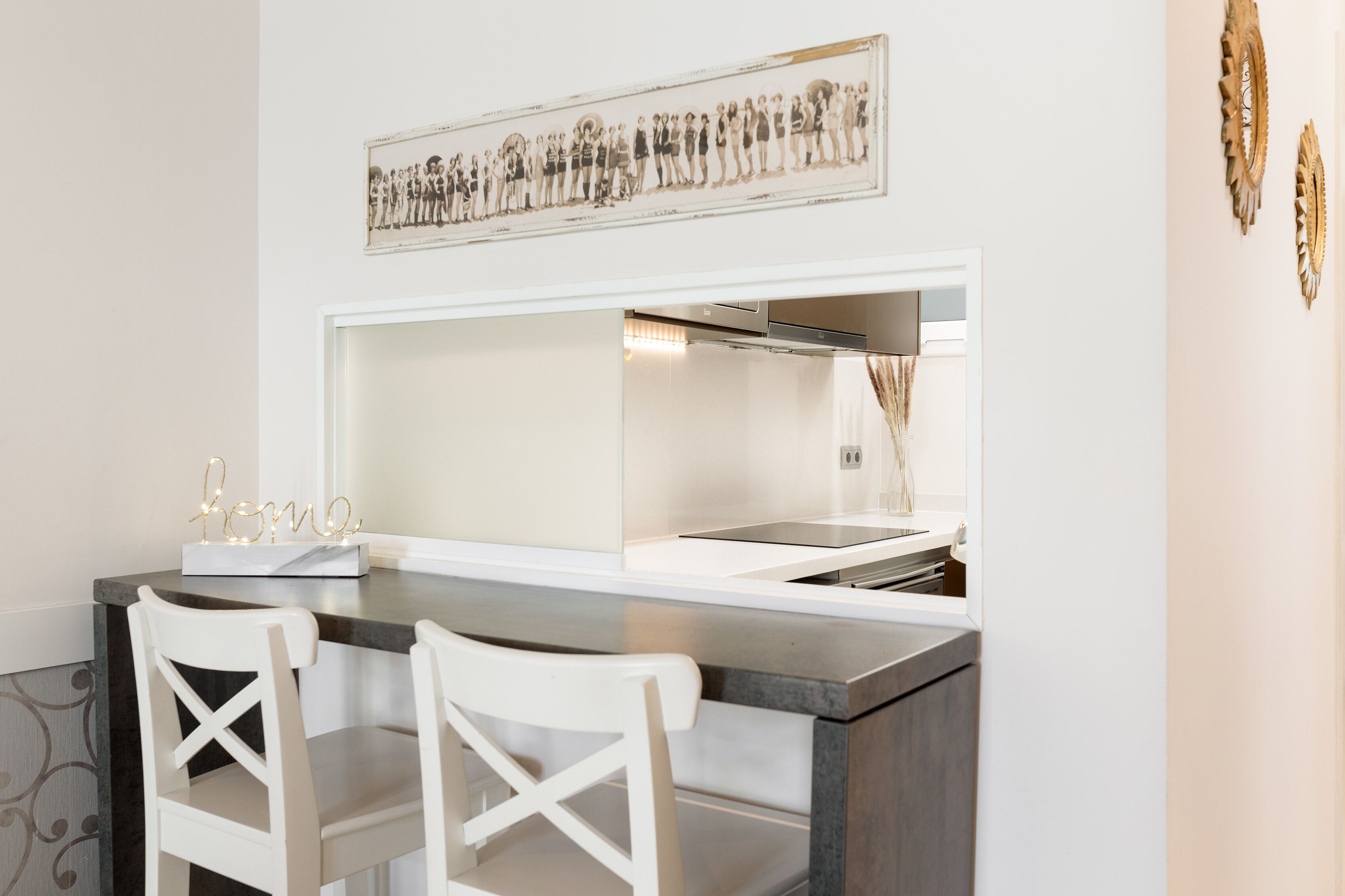 A modern kitchen with a white countertop, two white chairs, and a decorative light fixture spelling "home".