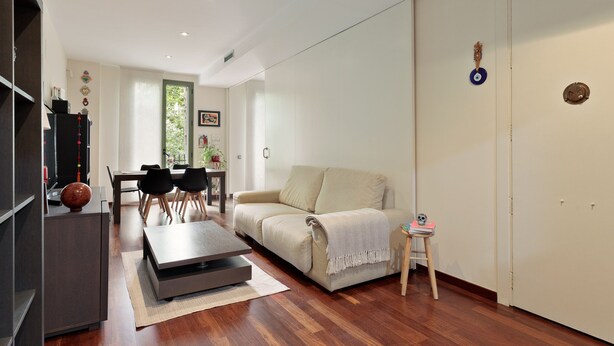 A modern living room with a sofa, dining table, and chairs. There is a small table with a decorative item and a stool. - Frida by EnjoyApartments (Barcelona)