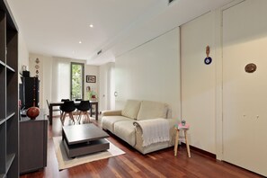 Ein modernes Wohnzimmer mit Sofa, Esstisch und Stühlen. Ein kleiner Tisch mit einem dekorativen Gegenstand und ein Hocker sind ebenfalls vorhanden.