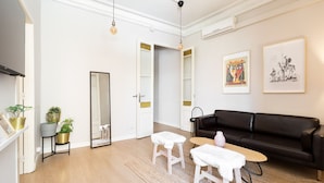 Een ruime woonkamer met een zwarte leren bank, twee witte krukken, een klein rond tafeltje en een grote spiegel. Er hangen twee kunstwerken in lijsten aan de muur en op een plank staat een pot met een plant.