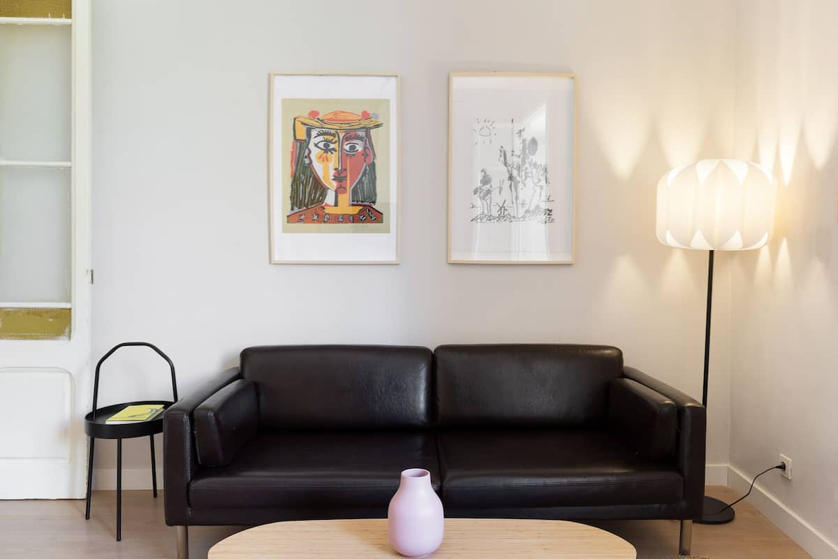 A modern living room with a black leather sofa, a small round table with a vase, and two framed artworks on the wall.