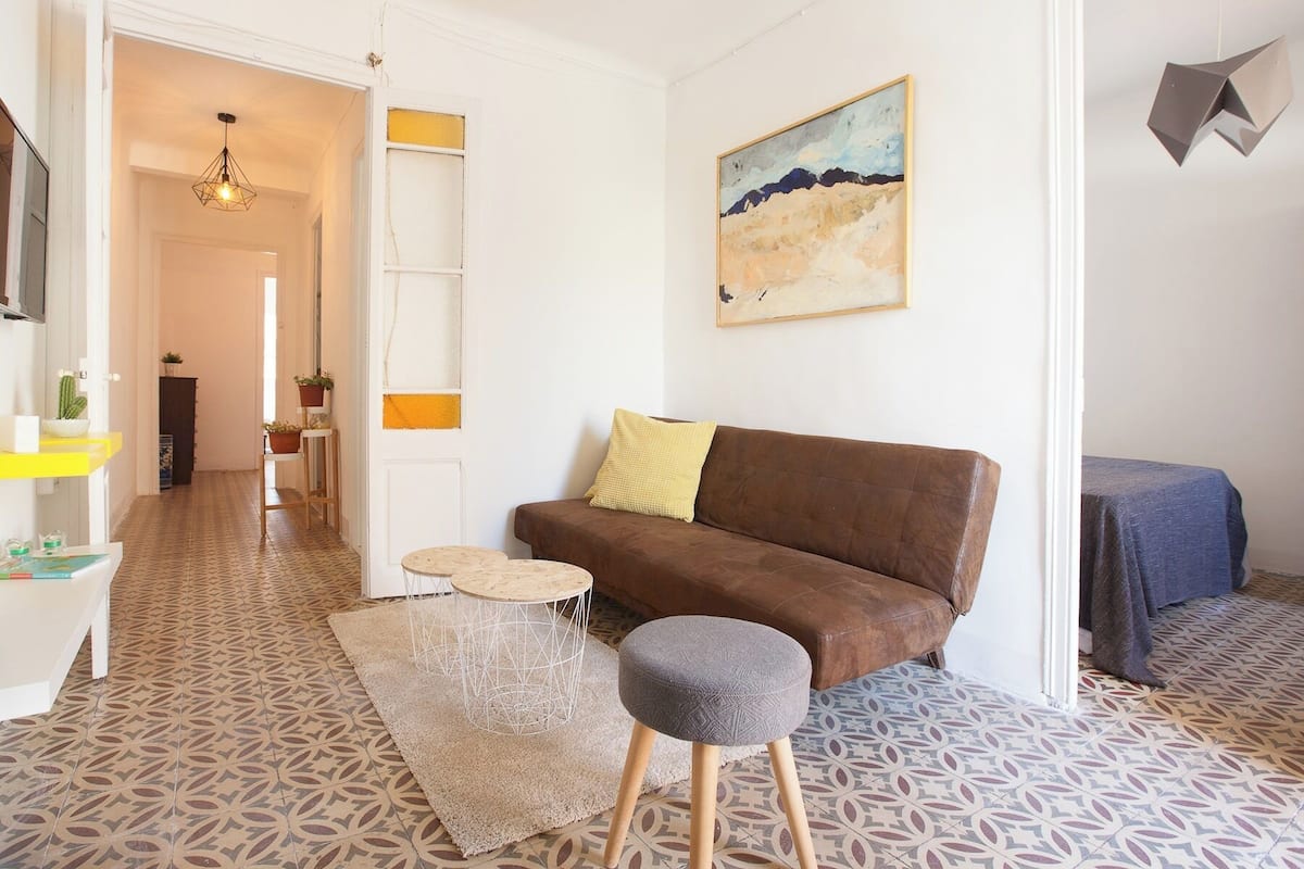 A living room with a brown sofa, a round grey ottoman, a small white table, and a patterned floor.
