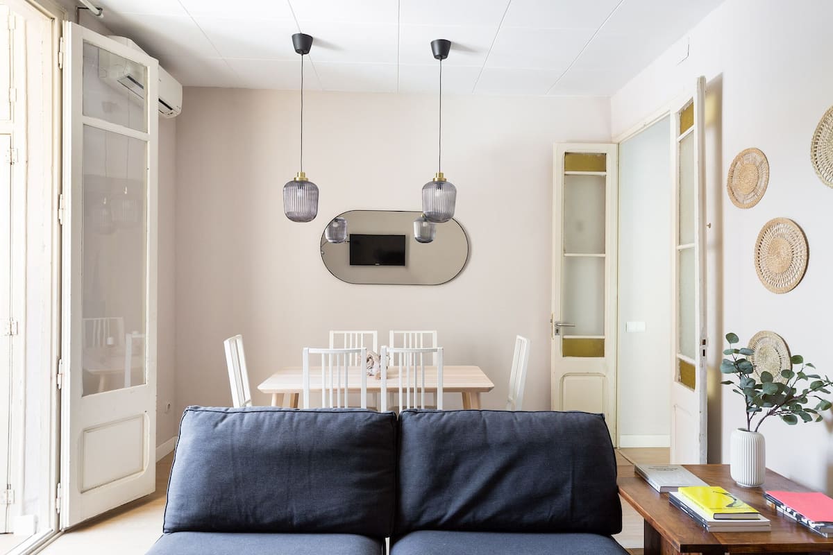 A modern living room with a sofa, a dining table with chairs, a TV mounted on the wall, and a bookshelf.