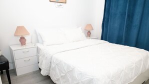 A neatly made bed with white bedding, two bedside tables with lamps, and a blue curtain. - Christon Exotic Suite - A Homey Suite (Halifax)