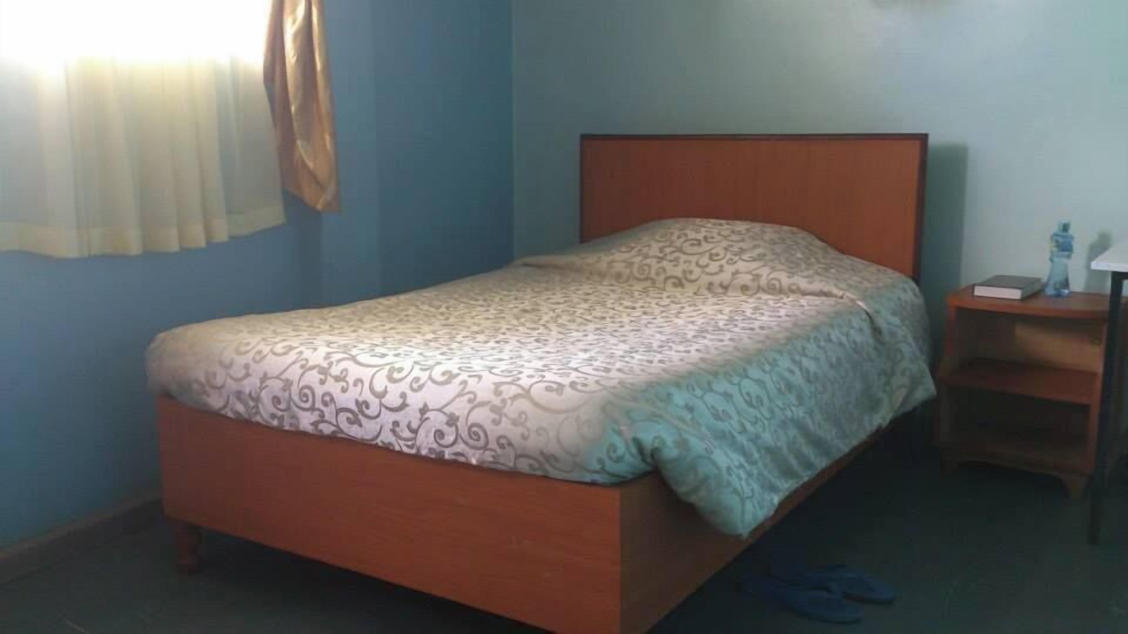 A single bed with a patterned bedspread, a wooden headboard, and a nightstand with a lamp and a book.