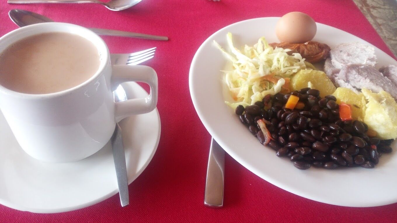 Una taza blanca de café sobre un platito, con un tenedor y una cuchara a un lado, y un plato con frijoles negros, col rallada, un huevo duro y rebanadas de carne.