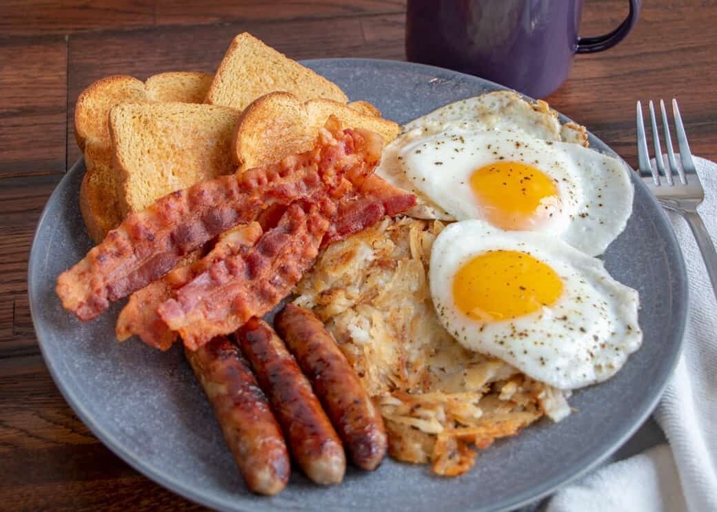 Un plato con tostadas, tocino, salchichas y huevos fritos.