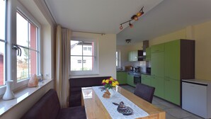 Un coin repas avec une table dressée pour deux personnes, un vase rempli de fleurs et, au fond, une vue sur la cuisine.