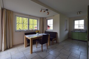 Ein Essbereich mit einem Holztisch, schwarzen Stühlen und einer Vase mit gelben Blumen. Große Fenster mit Vorhängen, eine grüne Küchenzeile und eine Deckenleuchte sind ebenfalls vorhanden.
