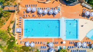 Vista aérea de un resort con dos piscinas grandes, tumbonas y sombrillas.