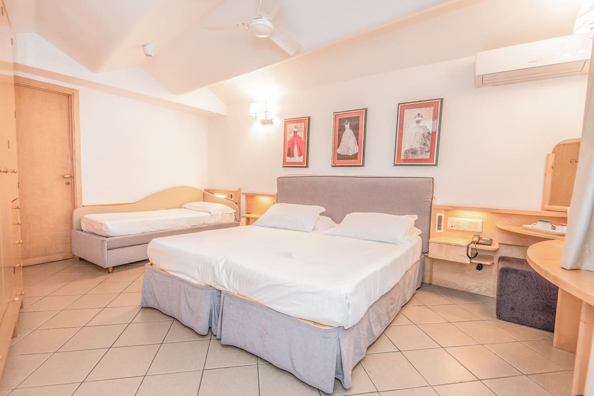 A hotel room with a large bed, a smaller bed, a desk with a chair, a wardrobe, and a bathroom with a mirror and sink.