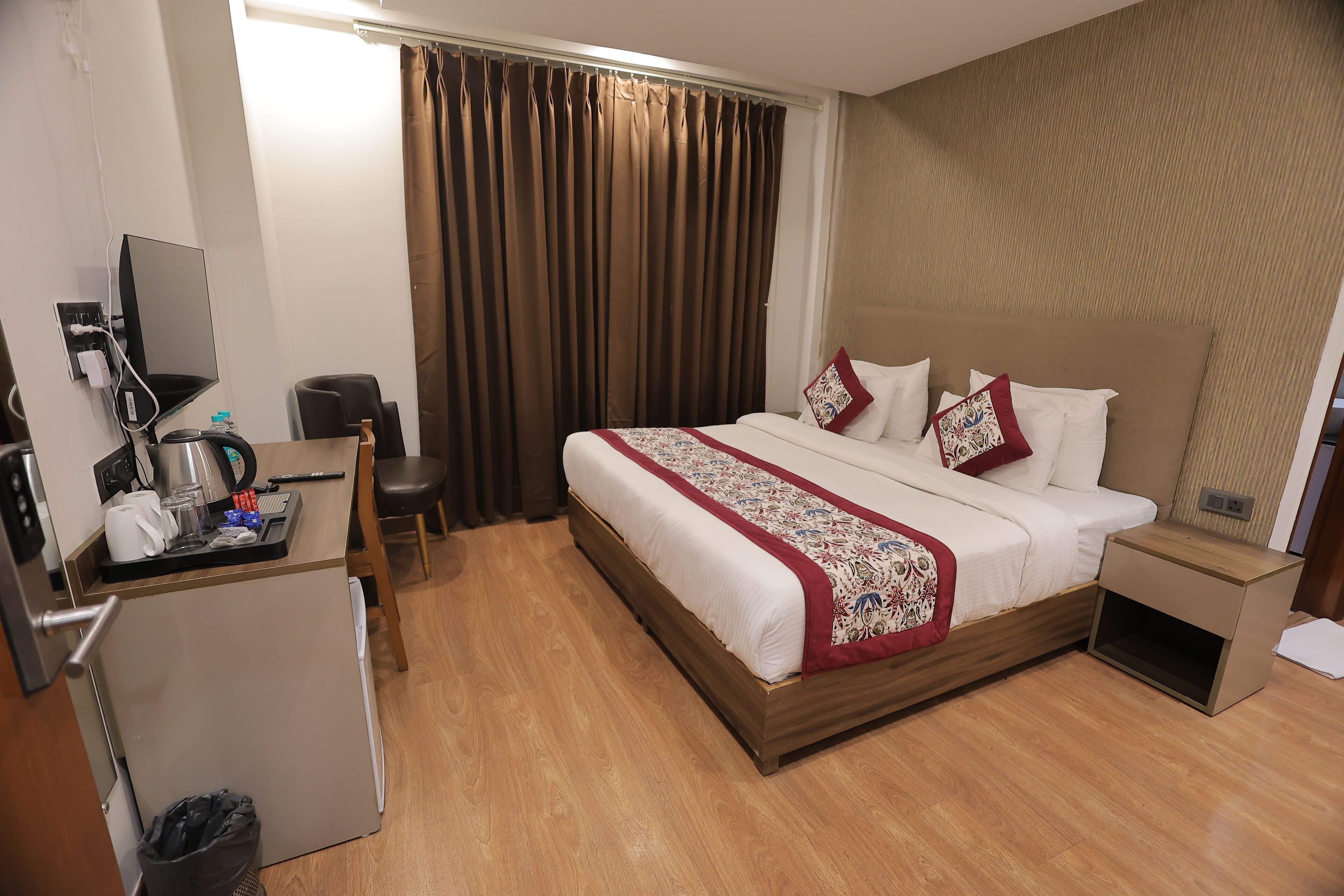 A hotel room with a bed, bedside table, desk with a computer, and a small trash bin.