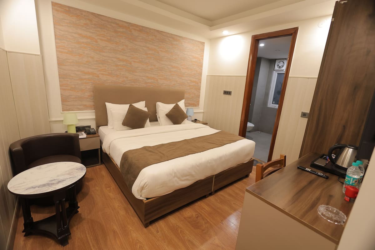 A hotel room with a bed, a round table, a chair, a nightstand, a lamp, a clock, and a door leading to another room.