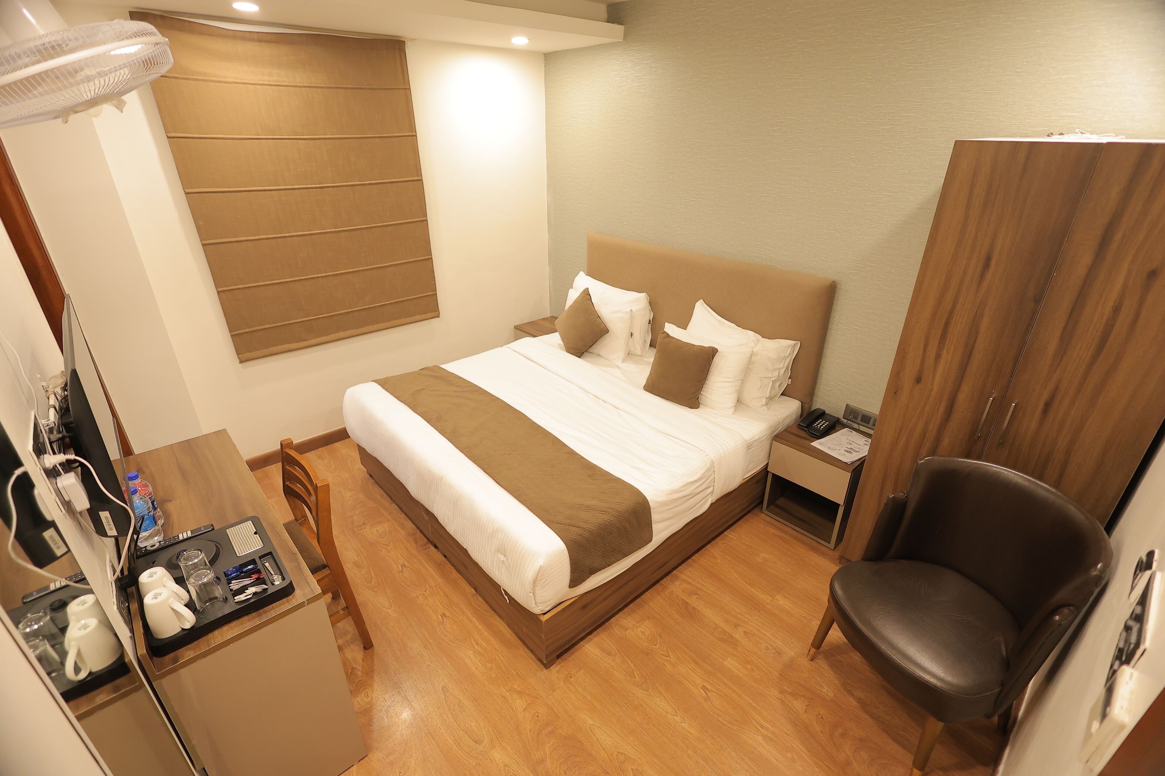 A neatly arranged hotel room with a bed, bedside table, chair, and a desk with a fan and various items.
