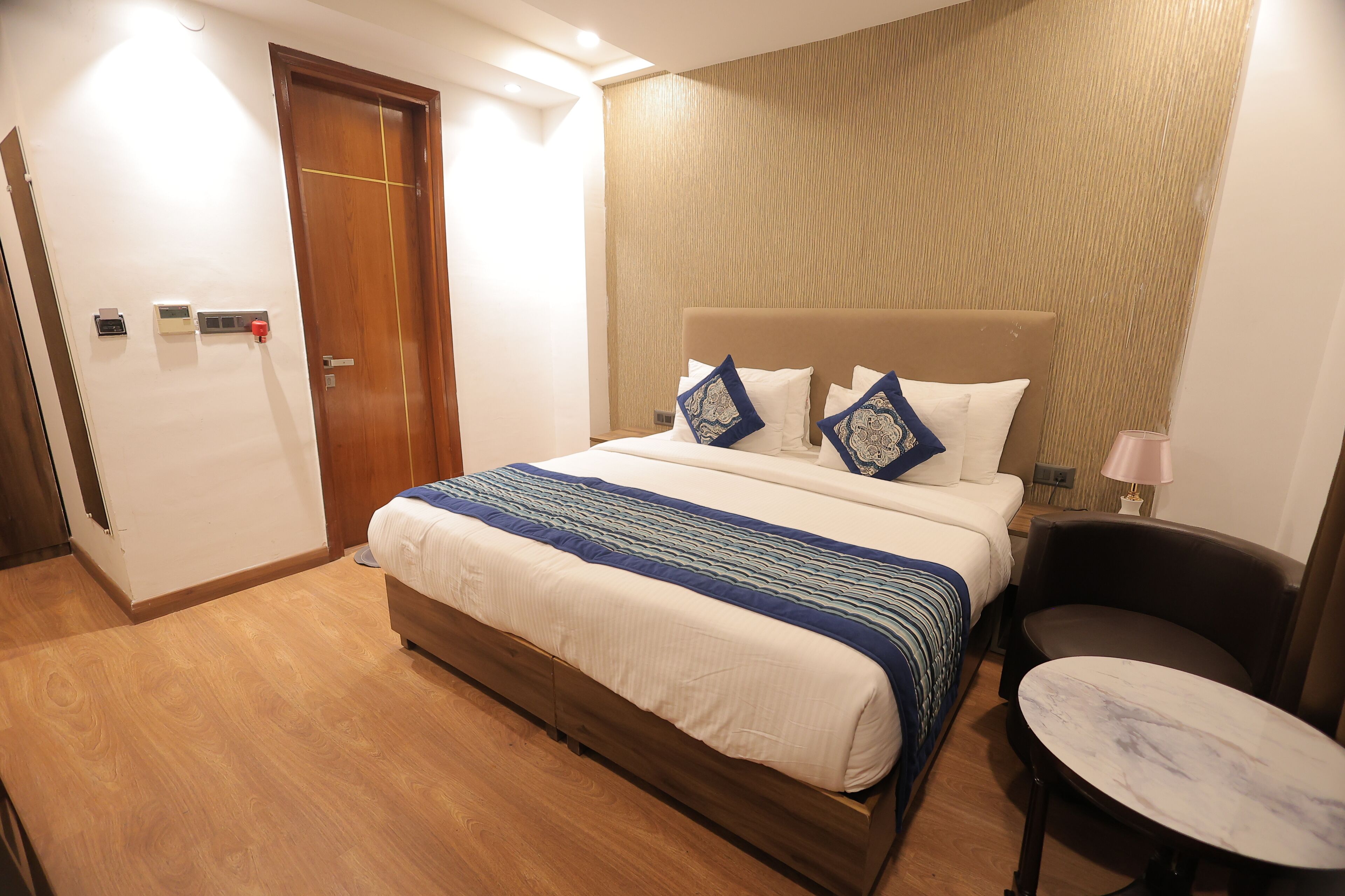 Una habitación de hotel con piso de madera, una cama con cabecera color beige, dos almohadas, una mesita de noche y una lámpara.