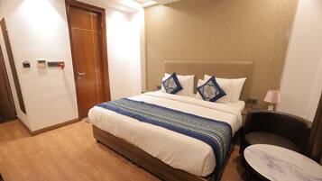 Una habitación de hotel con piso de madera, una cama con cabecera color beige, dos almohadas, una mesita de noche y una lámpara.