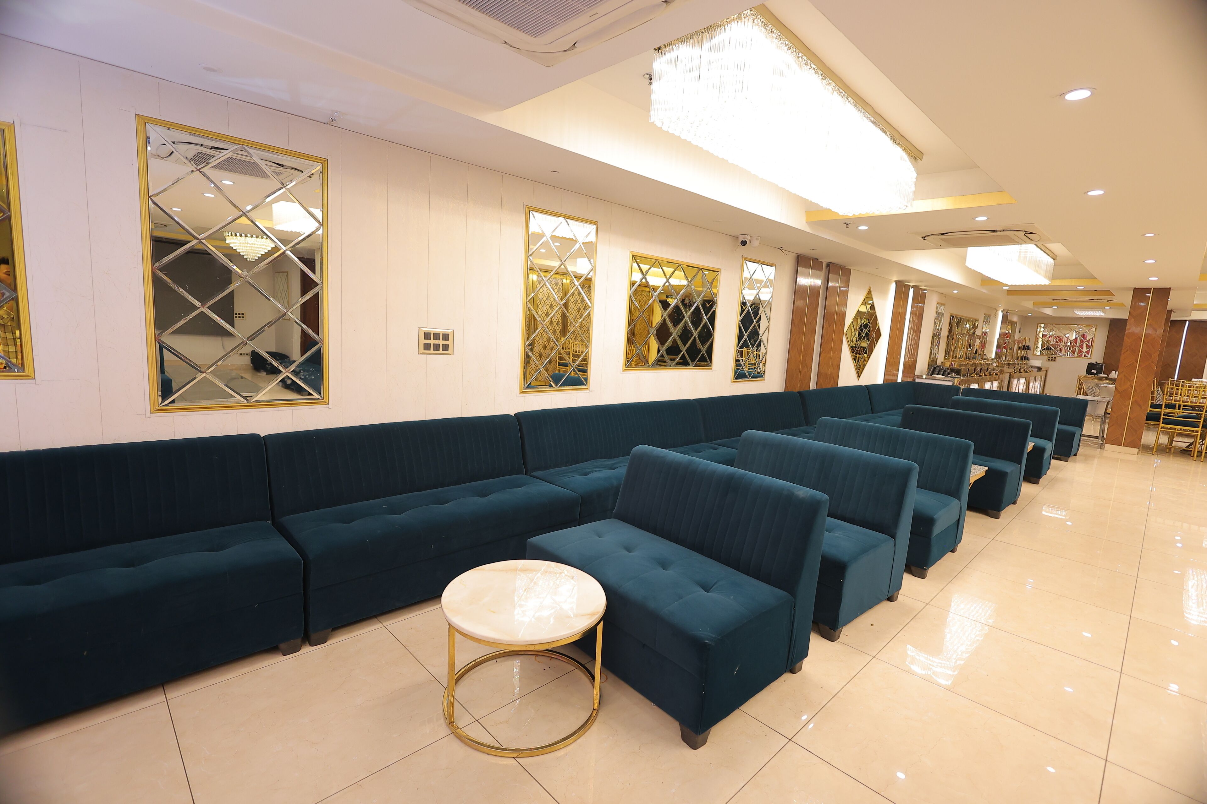 Un hall de hotel con sofás azul oscuro dispuestos en filas, una mesa redonda con marco dorado y espejos decorativos en las paredes.