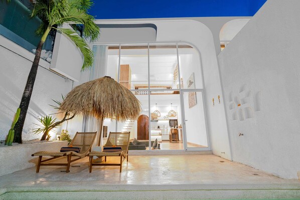 A modern, open-plan living area with a thatched-roof structure, lounge chairs, and a visible kitchen. - Villa Casa Zaya A (Canggu)