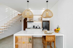 A modern kitchen with white cabinetry, a central island, and a dining area with wicker chairs. - Villa Casa Zaya A (Canggu)