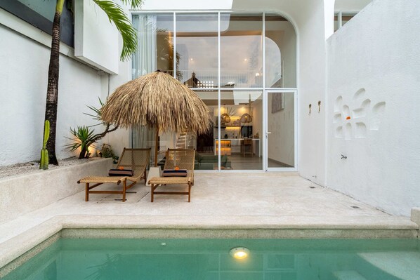 A poolside area with two lounge chairs and a thatched umbrella, a white wall with a decorative cut-out, and a glimpse of the interior through glass doors. - Villa Casa Zaya A (Canggu)