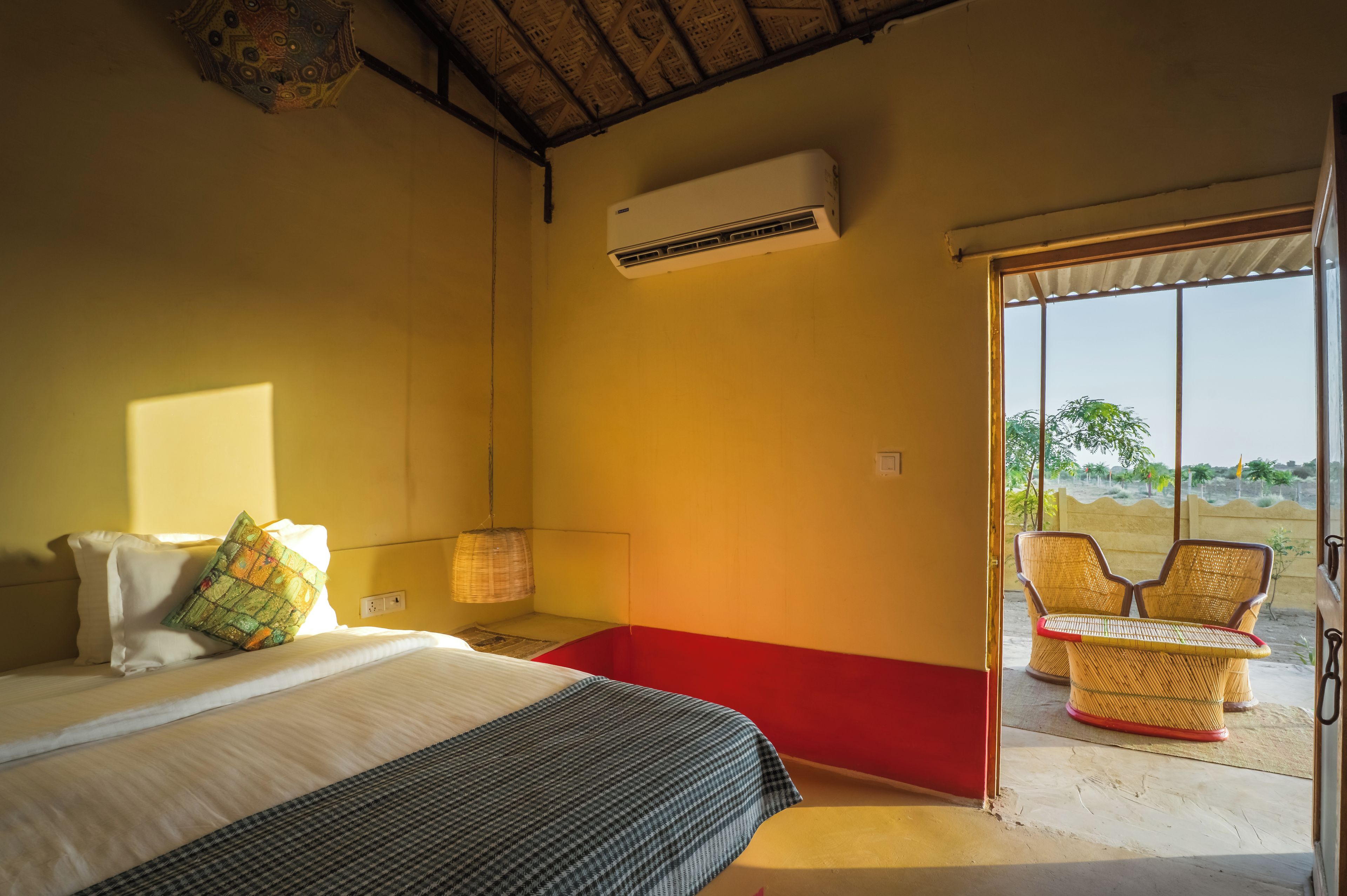 A room with a bed, a red bench, wicker chairs, and a large window.