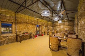 A spacious dining area with brick walls, multiple tables, wicker chairs, and ceiling fans. - Vijay Bagh Thar Village Resort (Jaisalmer)