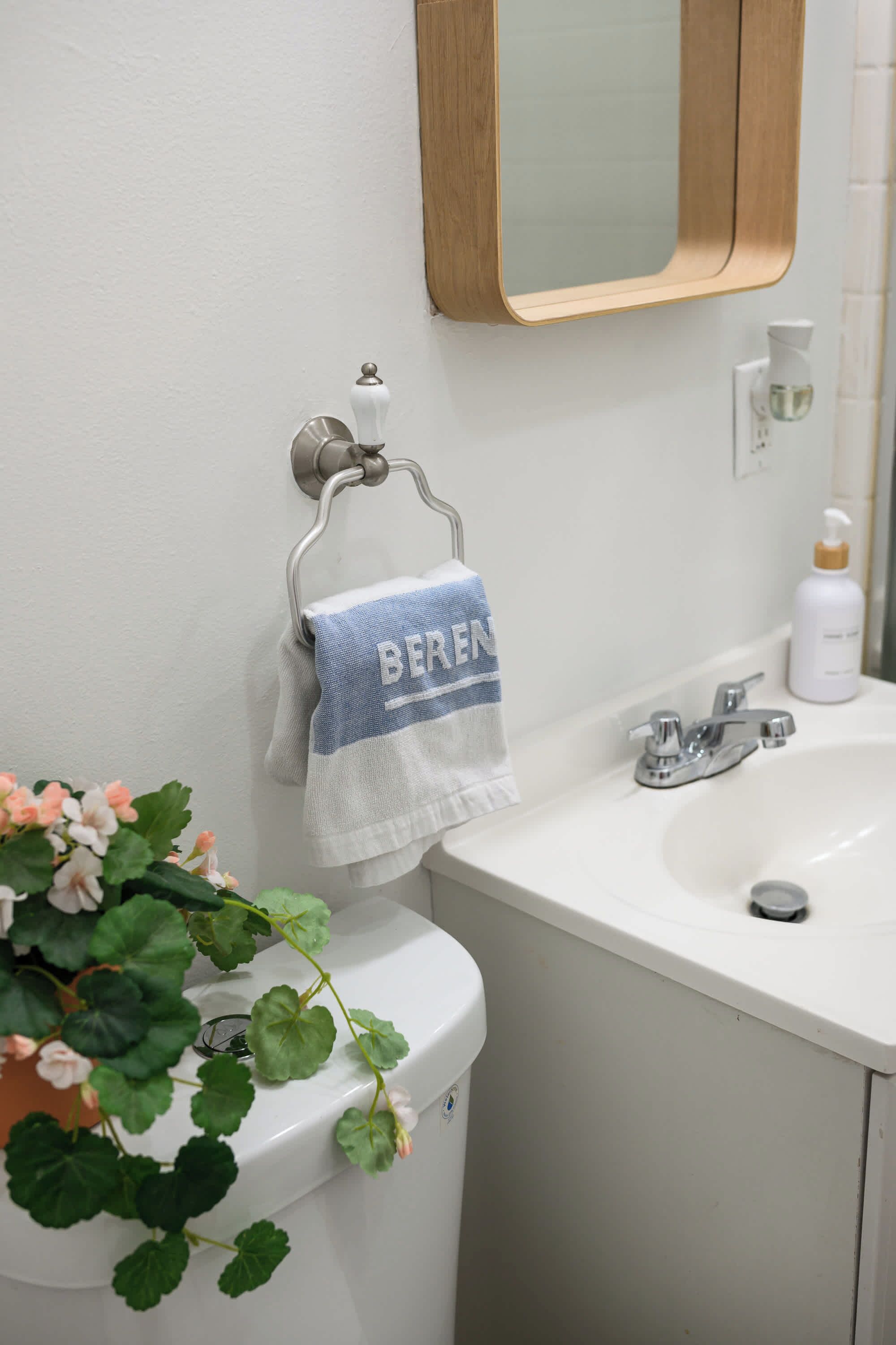 Ein Badezimmer mit einem weißen Waschbecken, einem Spiegel und einer Toilette. Ein Handtuch mit dem Schriftzug 'BEREN' hängt über der Toilette.