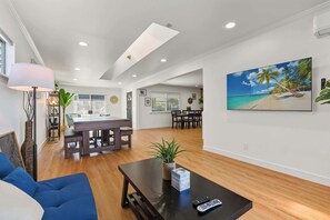 A spacious living area with a dining table, a coffee table, and a large painting of a beach scene. - Spacious 4BR House Free Parking w Backyard BBQ (La Puente)