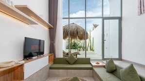 A modern living room with a green sofa, a flat-screen TV, and a large window offering a view of a patio with a thatched umbrella and cacti. - Villa Casa Zaya B (Canggu)
